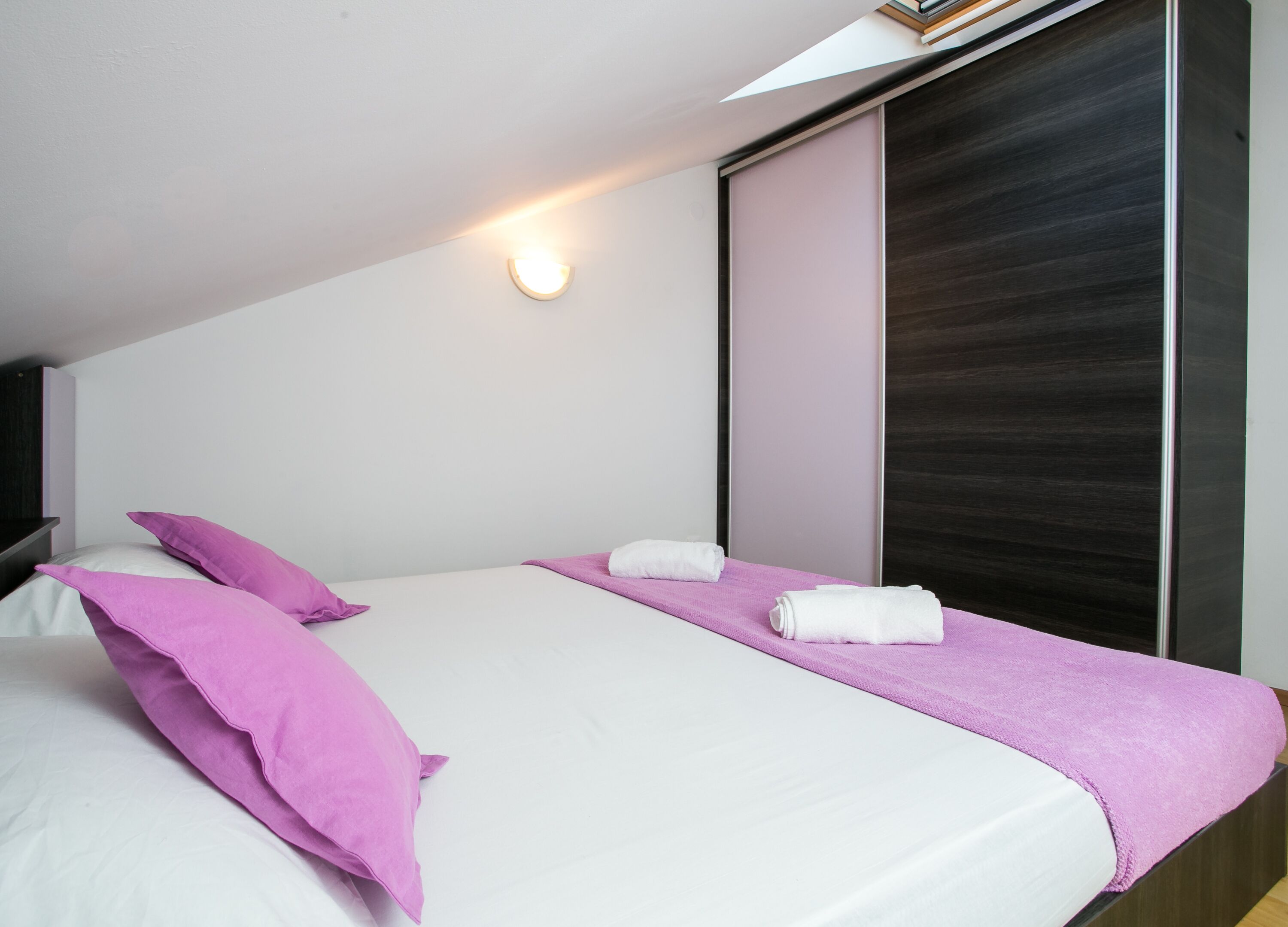 A modern bedroom with a single bed, two pillows, a pink towel, and a large window with a dark blind.