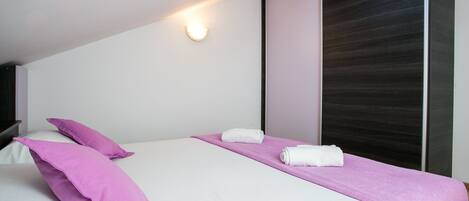 A modern bedroom with a single bed, two pillows, a pink towel, and a large window with a dark blind.