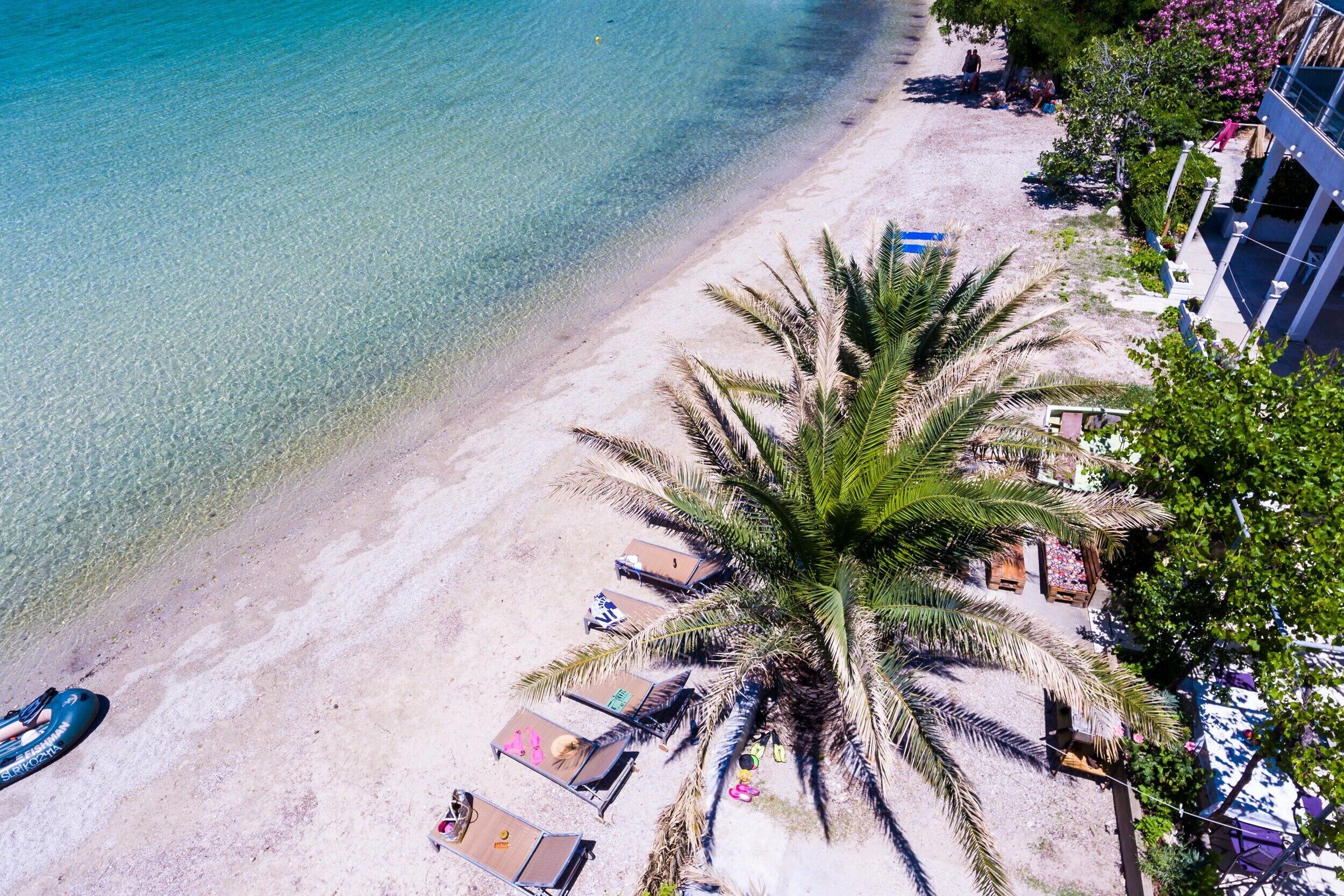 Ein Strand mit klarem türkisblauem Wasser, einem sandigen Ufer und einer Palme. Liegestühle und ein kleines Gebäude mit blauem Dach sind ebenfalls zu sehen.