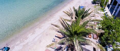 Ein Strand mit klarem türkisblauem Wasser, einem sandigen Ufer und einer Palme. Liegestühle und ein kleines Gebäude mit blauem Dach sind ebenfalls zu sehen.