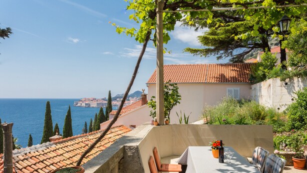 A terrace with a table and chairs, overlooking a coastal view with buildings and cypress trees.