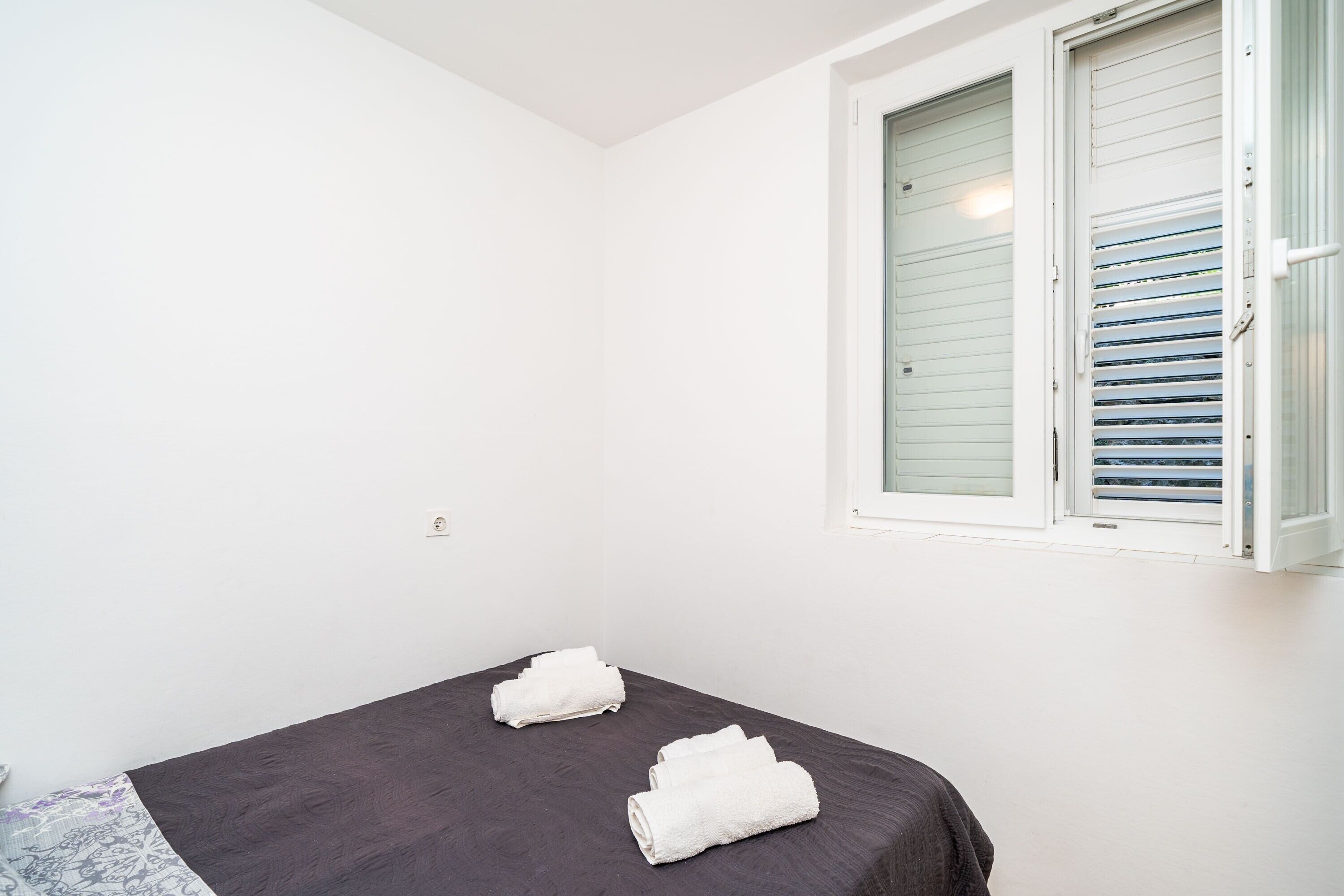 A small, single-bed room with a dark-colored bedspread, white towels, and a window with closed blinds.