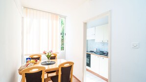Et lyst spiseområde med et bord og stole, et vindue med persienner, og udsigt til et køkken gennem en åben dør.
