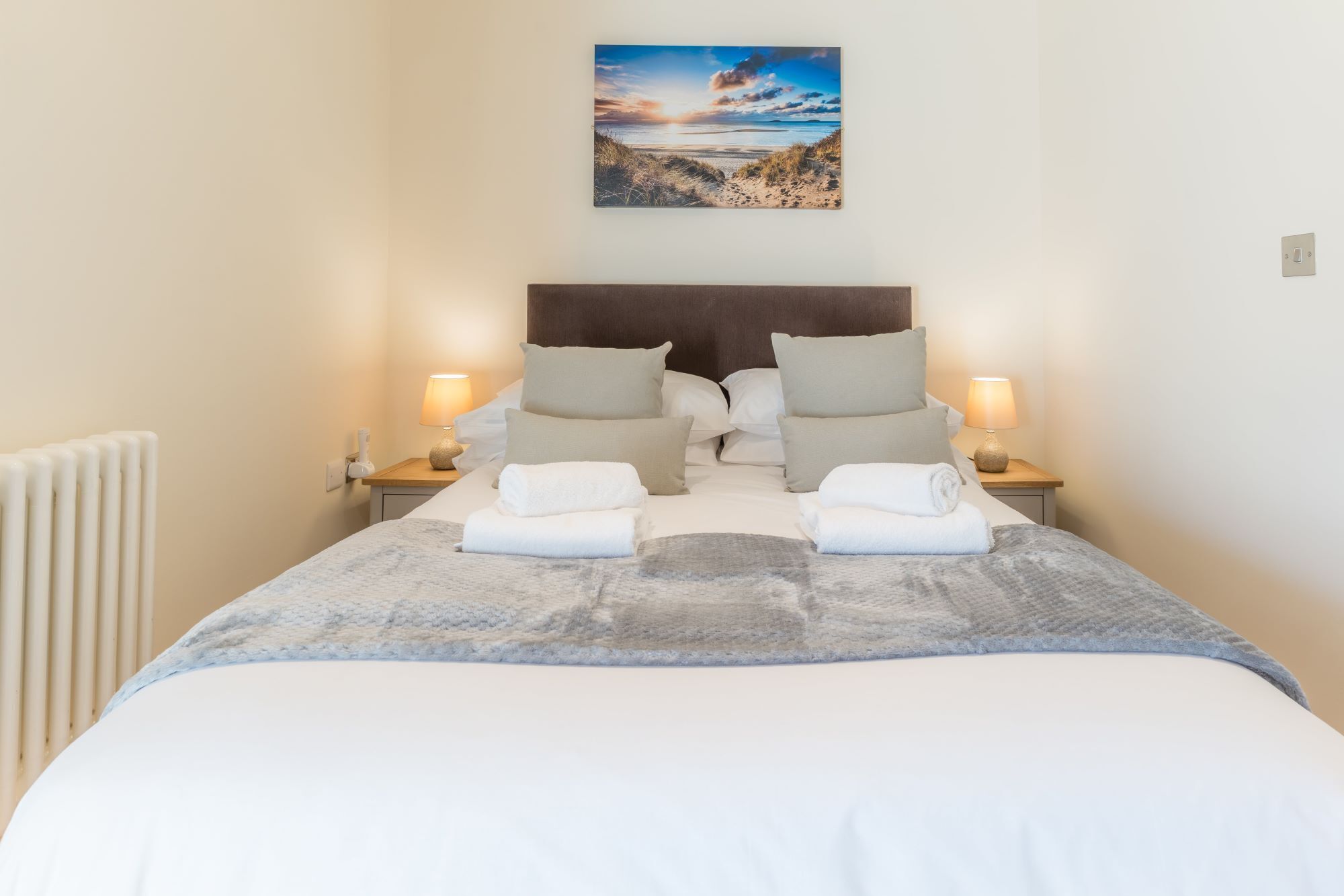 A neatly made bed with a headboard, two bedside lamps, and a painting of a beach scene above it.