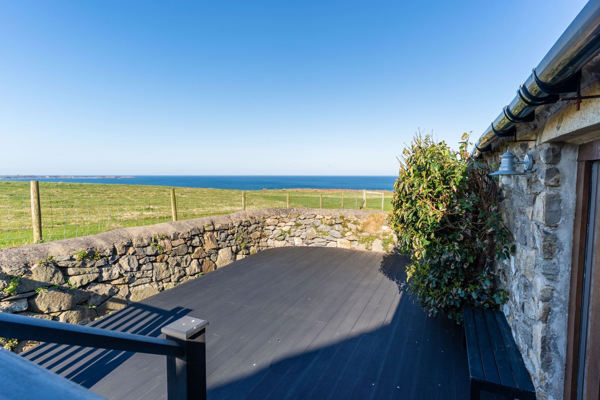 Un mur de pierre offrant une vue sur la mer, une terrasse en bois et un bâtiment en pierre surmonté d’un toit de chaume.