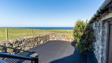 Un mur de pierre offrant une vue sur la mer, une terrasse en bois et un bâtiment en pierre surmonté d’un toit de chaume.