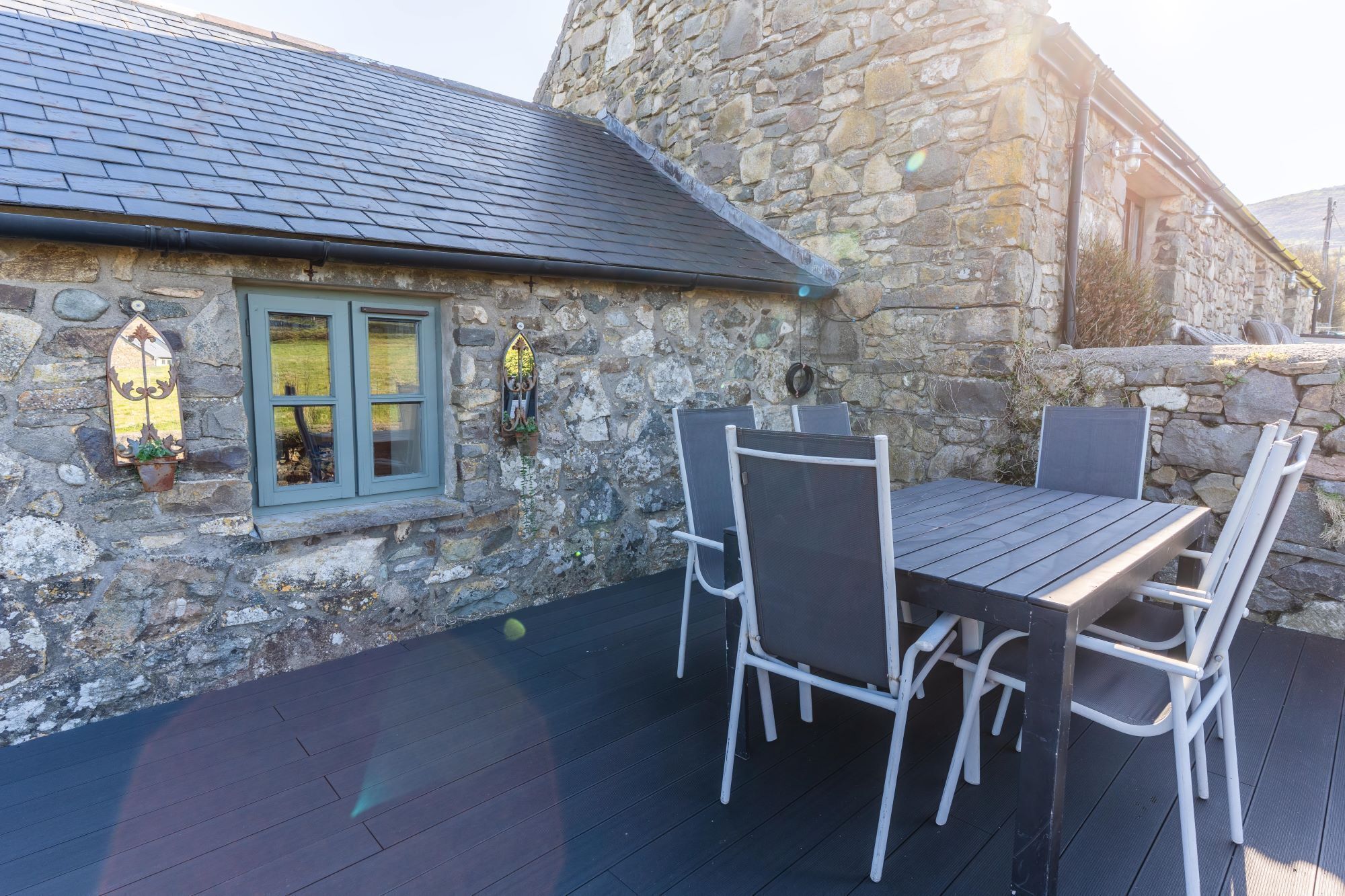 A stone building with a dark roof, a window with green shutters, and a table with chairs outside.