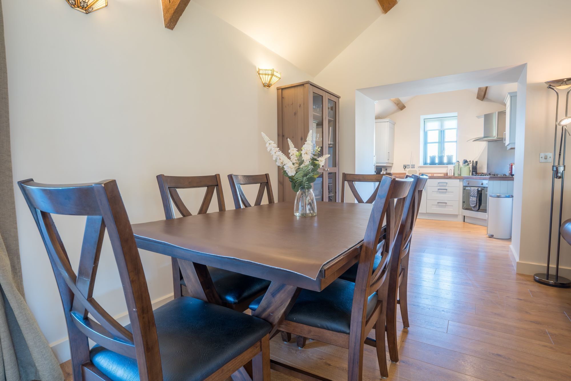 A dining area with a wooden table and chairs, a vase with flowers, and a kitchen in the background.