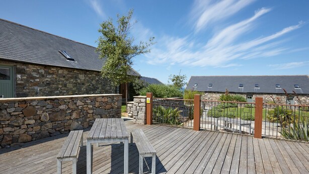 A wooden deck with a stone wall and a metal gate, leading to a stone building with a sloped roof. - The Tack Room (Pwllheli)