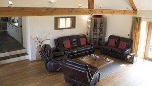 A spacious living room with a high ceiling and exposed wooden beams, furnished with leather sofas and armchairs, a coffee table, and a bookshelf. - The Tack Room (Pwllheli)