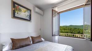 Un dormitorio con cama, un cuadro, aire acondicionado y un balcón con vistas a zonas verdes y colinas.