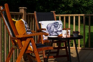 Two wooden deck chairs with cushions, a small table with a cup and saucer, and a railing. - Trewhiddle Villa 03 (St Austell)