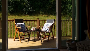 A deck with two wooden chairs and a small table, a glass door, and a view of a grassy area. - Trewhiddle Villa 03 (St Austell)