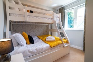 A bunk bed room with a geometric patterned headboard, a window with curtains, and a lamp on the left. - Trewhiddle Villa 03 (St Austell)