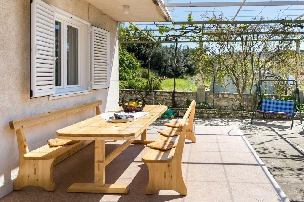 Mesa y bancos de madera al aire libre, con un muro de piedra y follaje en el fondo.