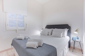 A neatly made bed with a grey headboard and white bedding, a bedside table with a lamp, and a window with a view of a snowy landscape. - Sweett | Rocafort Principal 4 (Barcelona)