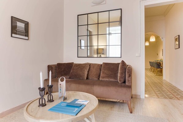 A living room with a brown sofa, a round wooden table with a blue folder, and a large mirror on the wall. - Sweett | Rocafort Principal 4 (Barcelona)