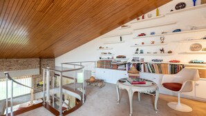 Une pièce de style « loft » avec un plafond en pente, un escalier en verre et un mur en briques blanches. On y trouve des étagères remplies de livres et d’objets décoratifs, une table ronde avec un livre rouge, et une chaise blanche avec un coussin rouge.