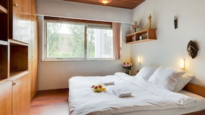 Une chambre avec un plafond en bois, une grande fenêtre, un lit recouvert de draps blancs et une petite étagère ornée d’objets décoratifs.