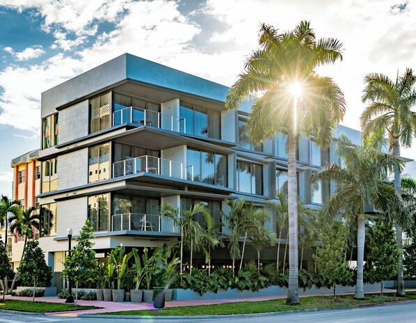 Un edificio moderno de varios pisos con balcones, amplias ventanas y exuberante jardinería, incluyendo palmeras.