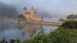 Un château historique doté de deux tours, situé sur la rive d’une rivière, dans une atmosphère brumeuse.