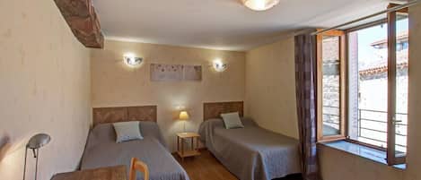 A room with two beds, a wooden table, a lamp, and a window with curtains.