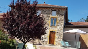 Una casa de pedra amb una porta i persianes de fusta, un petit jardí amb un arbre i dues cadires blanques, i una zona de pati amb taula i sombrilla.