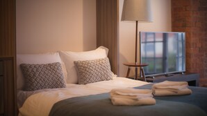 A neatly made bed with pillows, a lamp, and a folded towel on a bed in a room with a brick wall and a television. - Riverside Studios (York)