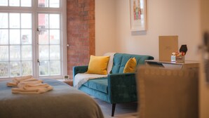 A hotel room with a bed, a blue armchair, a brick wall, a window, and a framed picture on the wall. - Riverside Studios (York)