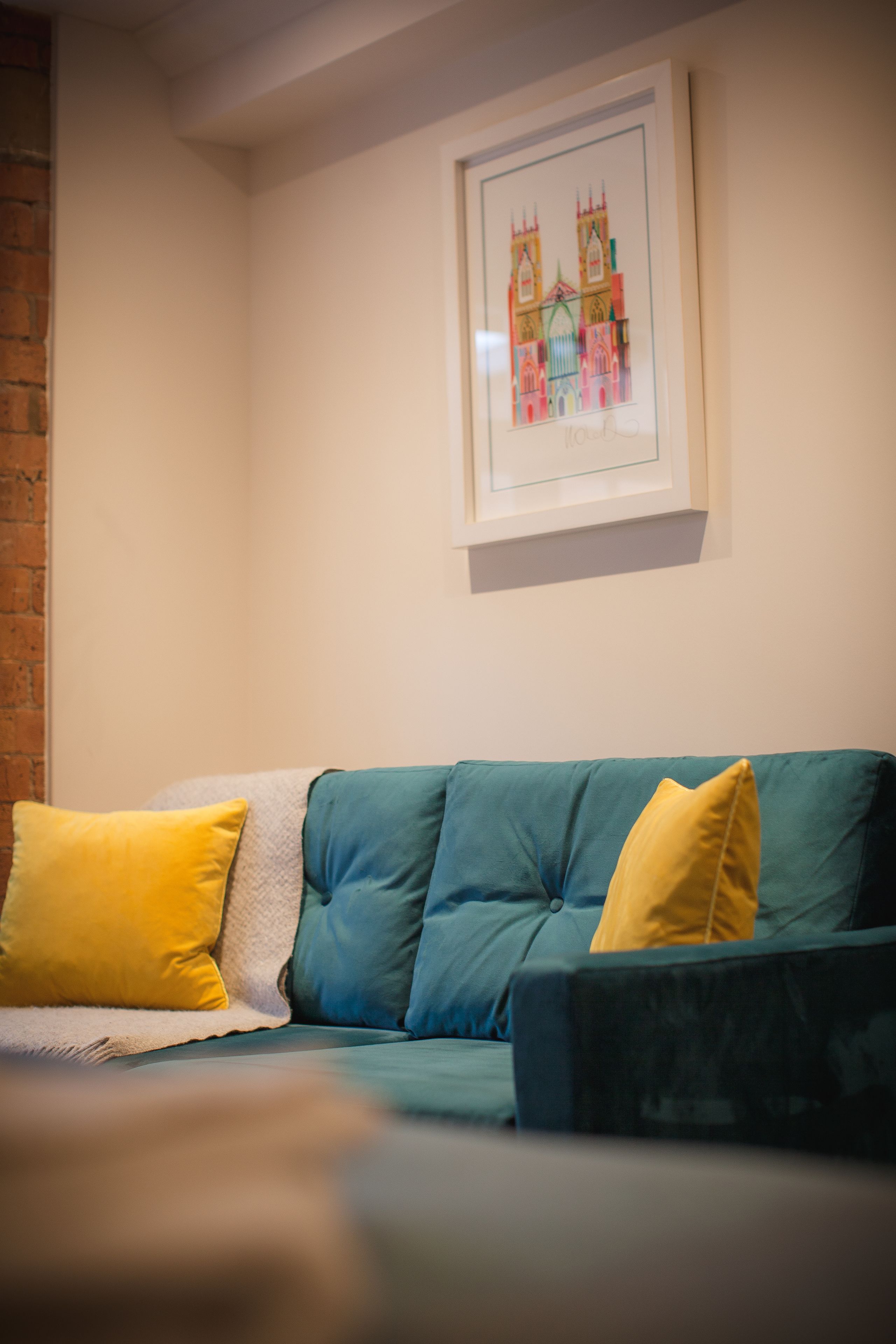 A green sofa with yellow and white cushions, a framed picture of a building on the wall, and a brick wall to the left.
