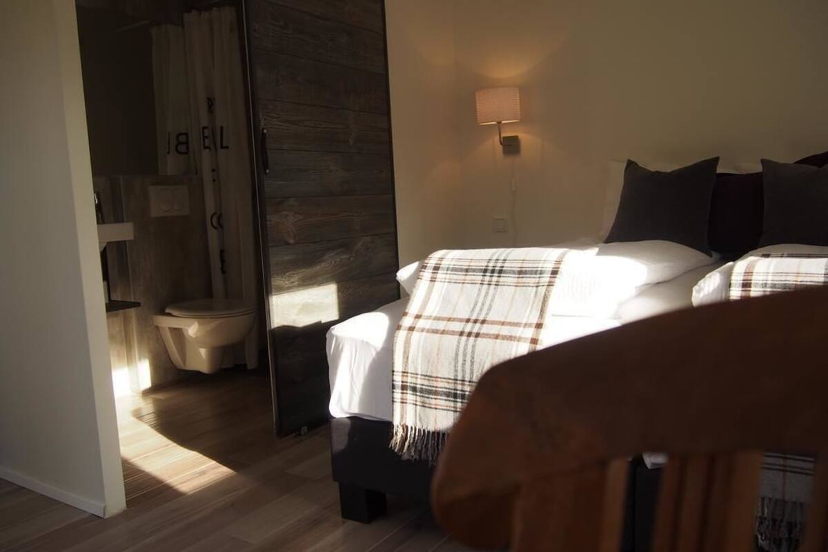 A bedroom with a large bed, a bedside lamp, and a view of a bathroom with a toilet and shower curtain.