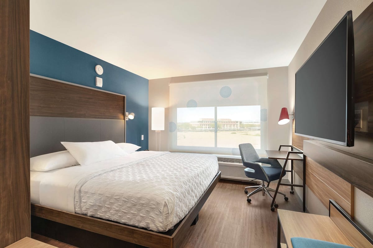 Quarto de hotel com cama, escrivaninha, cadeira, televisão e janela grande.