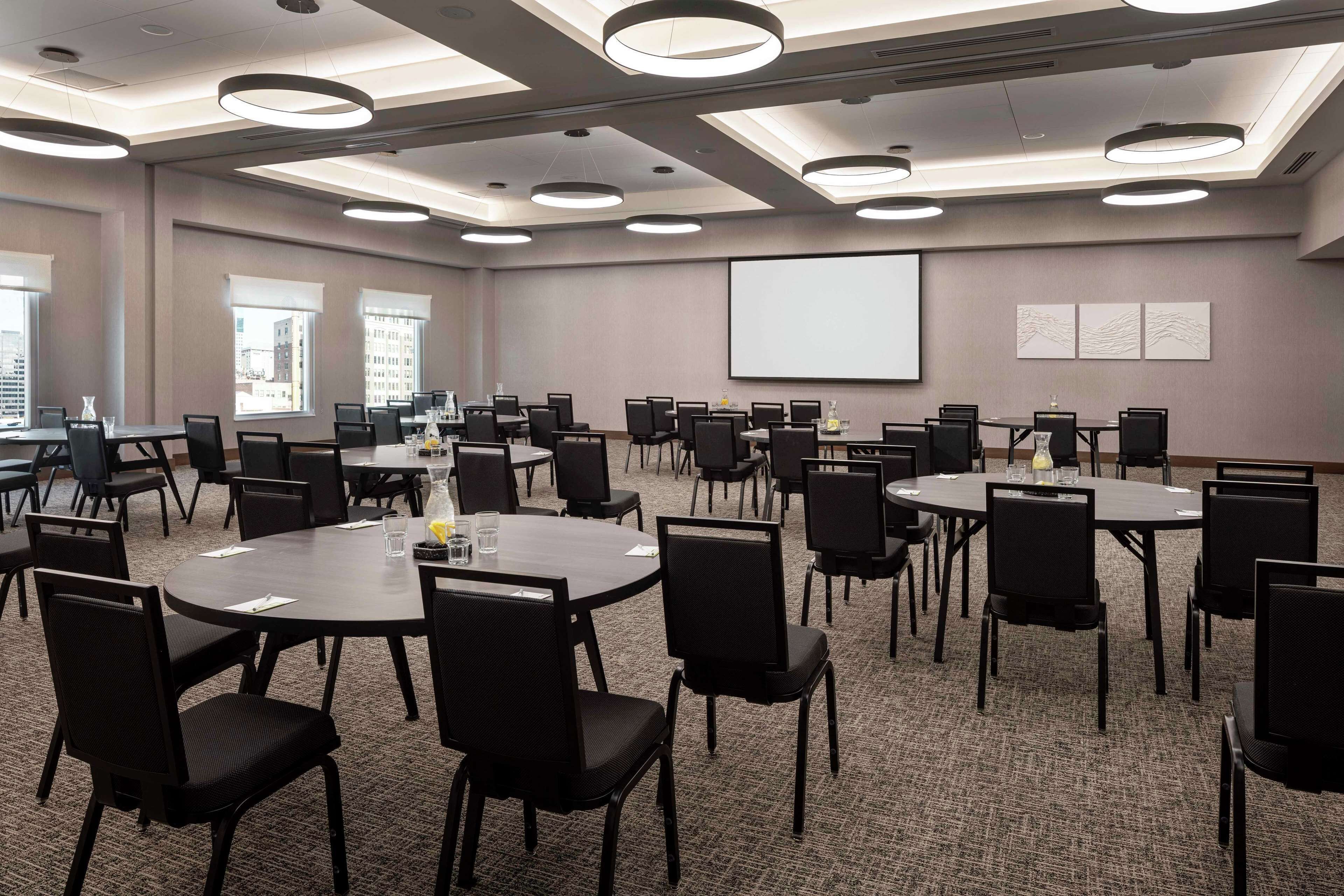 A conference room with multiple tables and chairs, a projector screen, and large windows with a city view.
