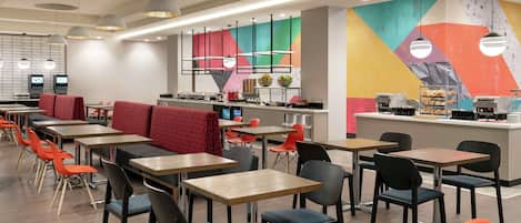 A modern restaurant with multiple tables and chairs, a counter with food items, and a colorful wall mural.