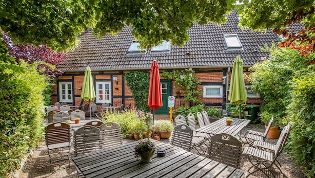 An outdoor patio area with wooden tables and chairs, surrounded by greenery and a brick building with a tiled roof. - Hotel Lindenkrug (Riepsdorf)