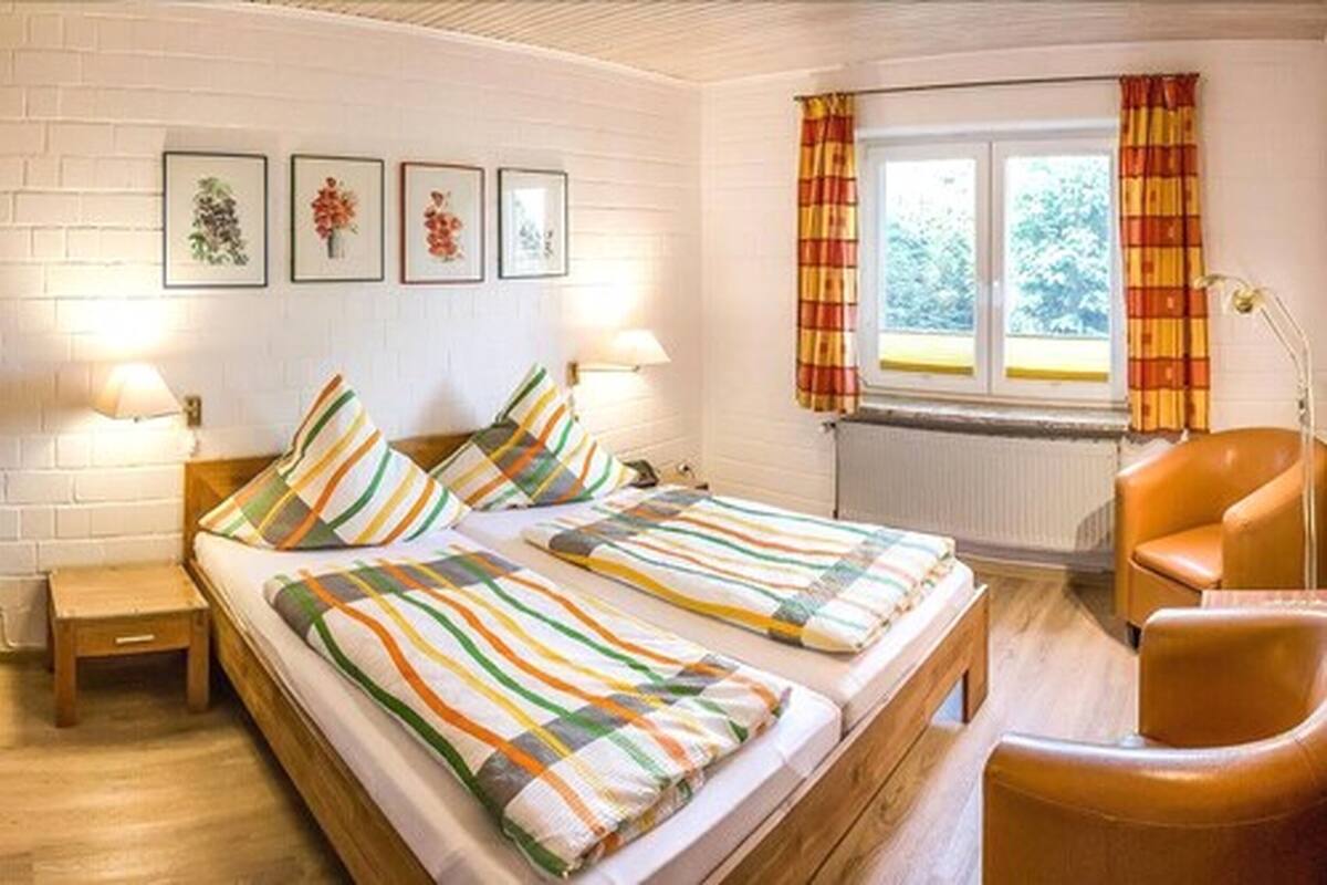 Una habitación con dos camas, una silla, una mesita y un ventanal con cortinas.