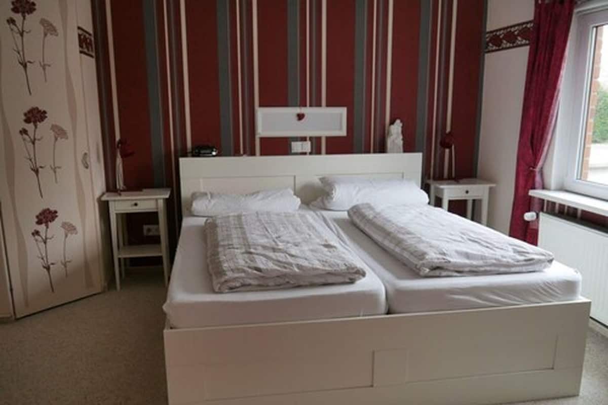 Un dormitorio con cama blanca, dos mesitas de noche, un tocador y un armario, junto a una ventana con cortinas.