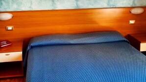 A bedroom with a wooden headboard, a bed with a blue bedspread, and a wall-mounted painting of a sunset with palm trees. - Hotel 2000 Fabriano (Fabriano)