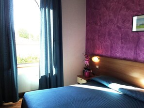 A hotel room with a bed, a nightstand, a purple wall, and a window with curtains. - Hotel 2000 Fabriano (Fabriano)