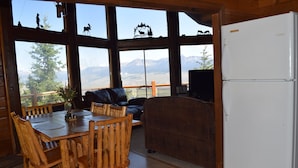 A wooden dining table with chairs, a sofa, and a white refrigerator in a room with large windows overlooking mountains. - Breath Taking Views-Near Yellowstone New Hot Tub.  Just off Union Pass! (Dubois)