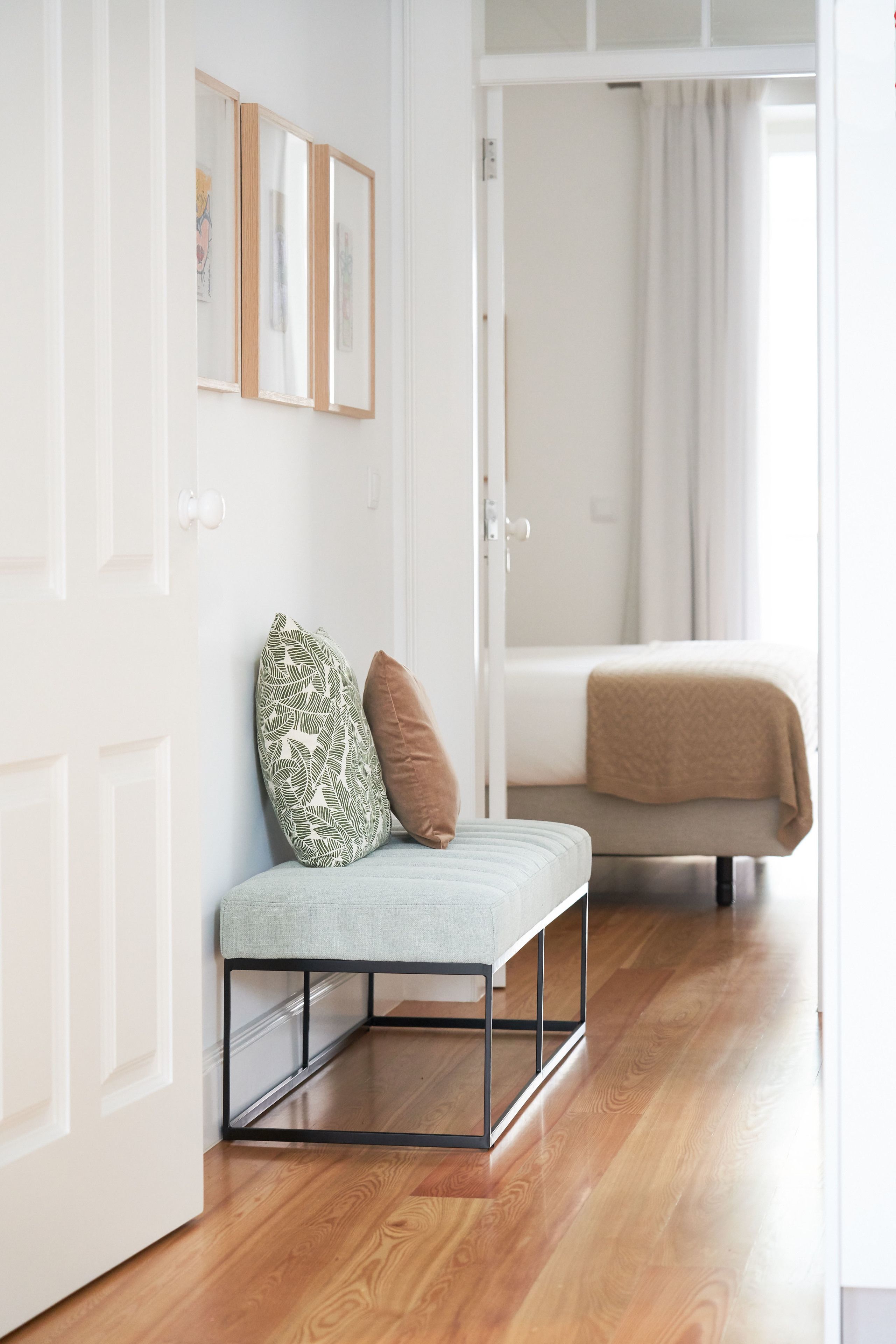 Une chambre moderne avec un plancher en bois, un banc agrémenté de coussins, un lit recouvert d’une couverture beige et des œuvres d’art encadrées accrochées au mur.