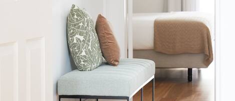 A modern room with a wooden floor, a bench with cushions, a bed with a beige blanket, and framed artwork on the wall.