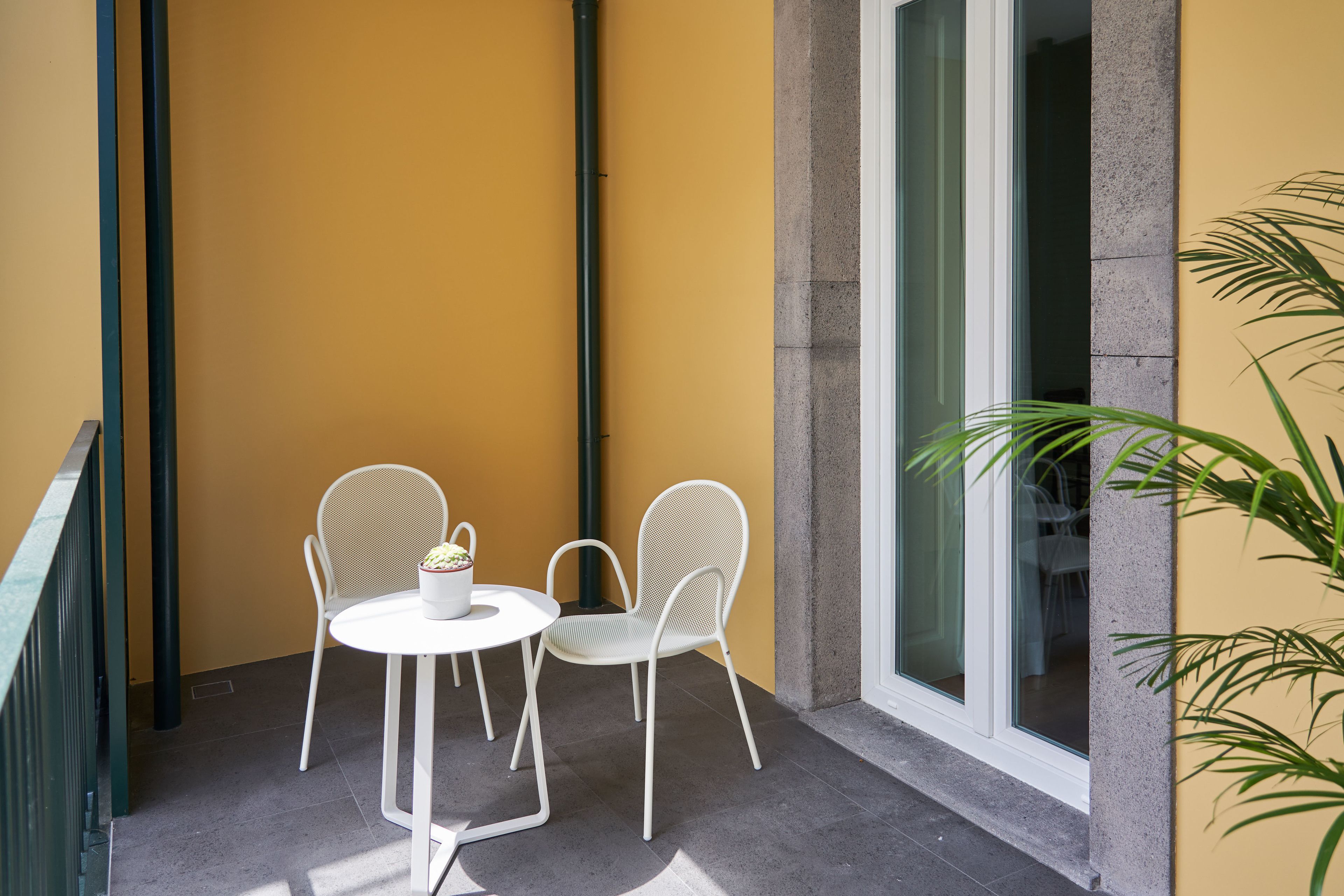 A balcony with two white chairs and a small table, a yellow wall, and a large window.