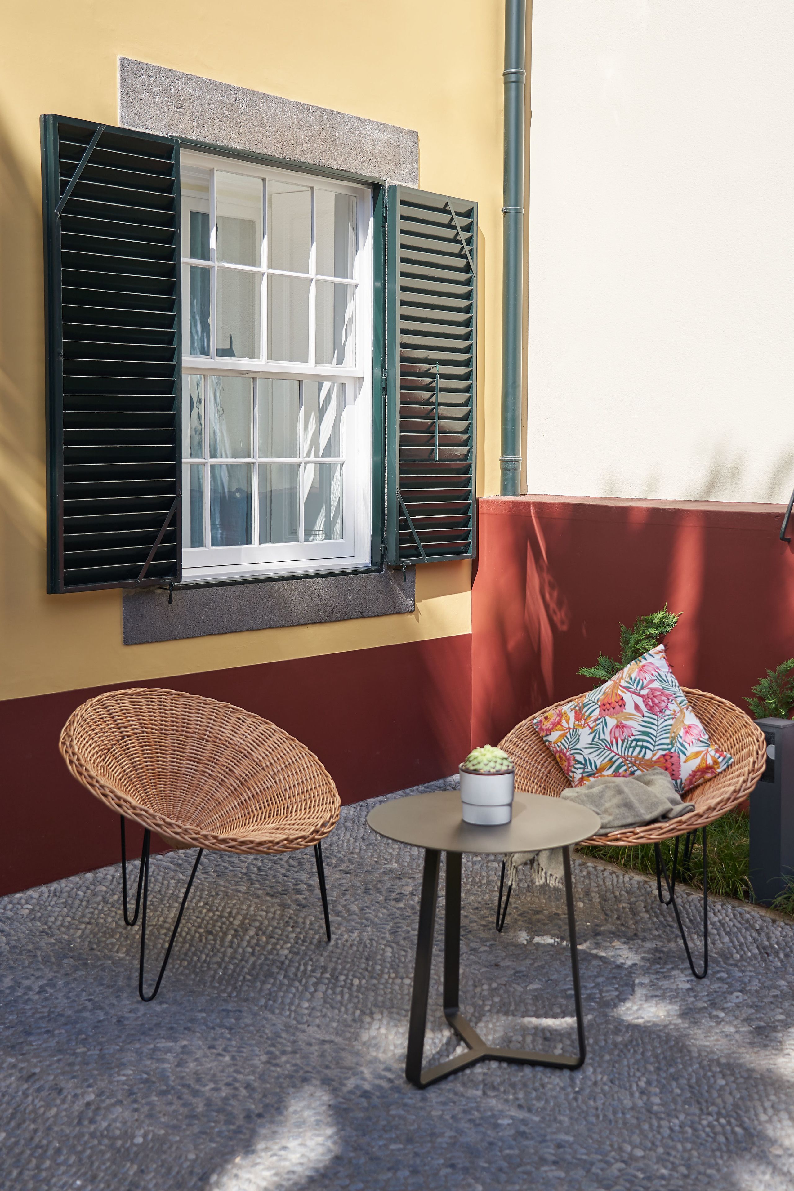 Ein kleiner Sitzbereich im Freien mit Korbstühlen und einem runden Tisch, ein Fenster mit Fensterläden und eine rote Wand.