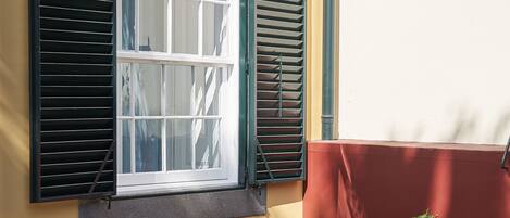 A small outdoor seating area with wicker chairs and a round table, a window with shutters, and a red wall.