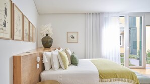 A modern bedroom with a wooden headboard, a large bed with white bedding and green pillows, a vase with white flowers, and a view of the outdoors through a glass door. - Good Memories Lodge (Funchal)