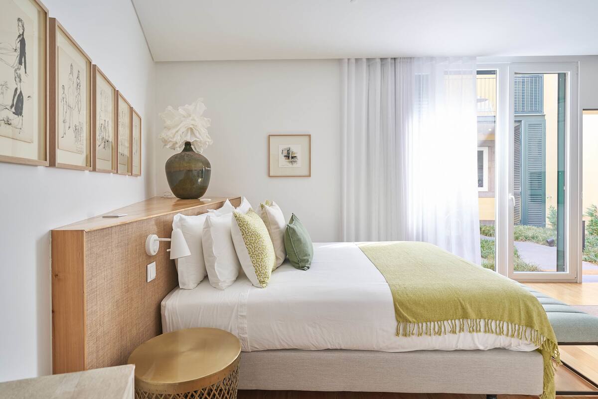 A modern bedroom with a wooden headboard, a large bed with white bedding and green pillows, a vase with white flowers, and a view of the outdoors through a glass door.