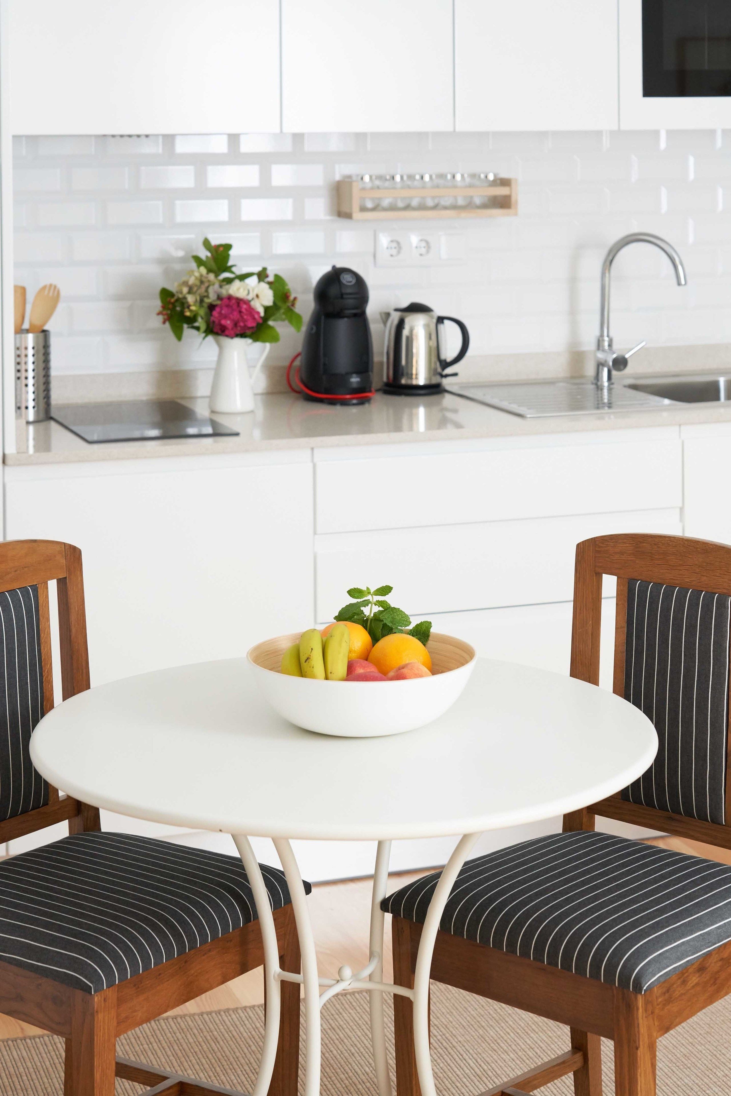 A modern kitchen with white cabinets, a white countertop, a coffee maker, a kettle, and a bowl of fruit on a round table.