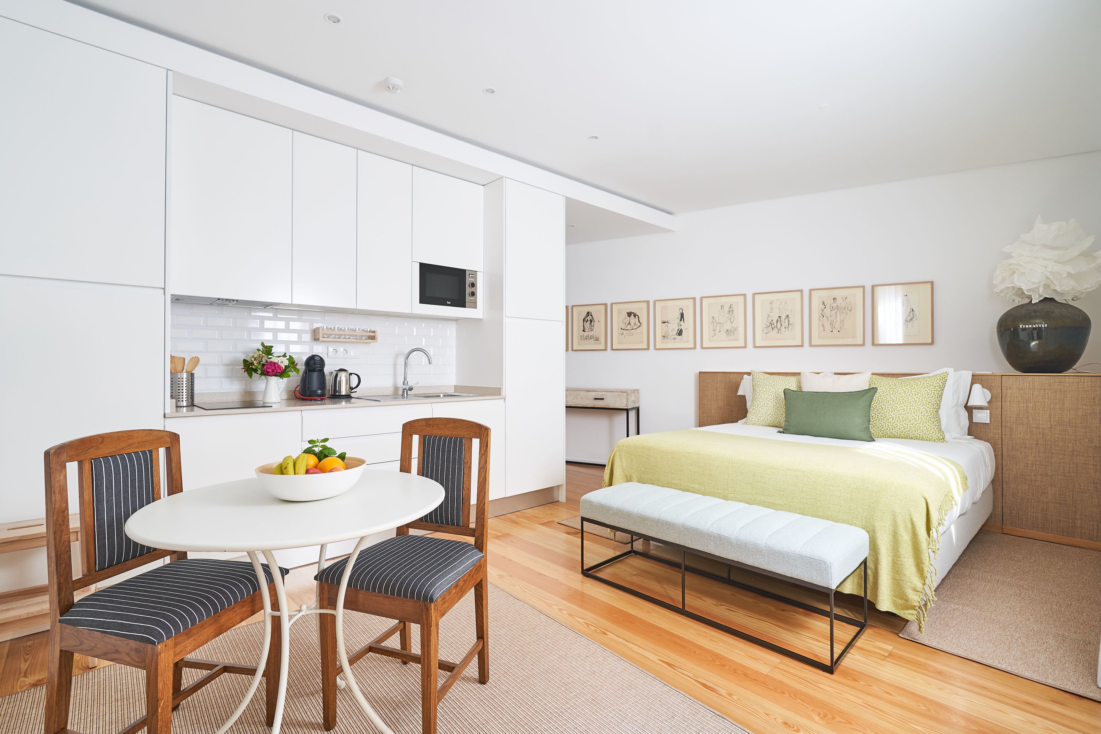 A modern, compact room with a kitchenette, a dining area with a table and chairs, a bed with a bench, and a wall with framed artwork.