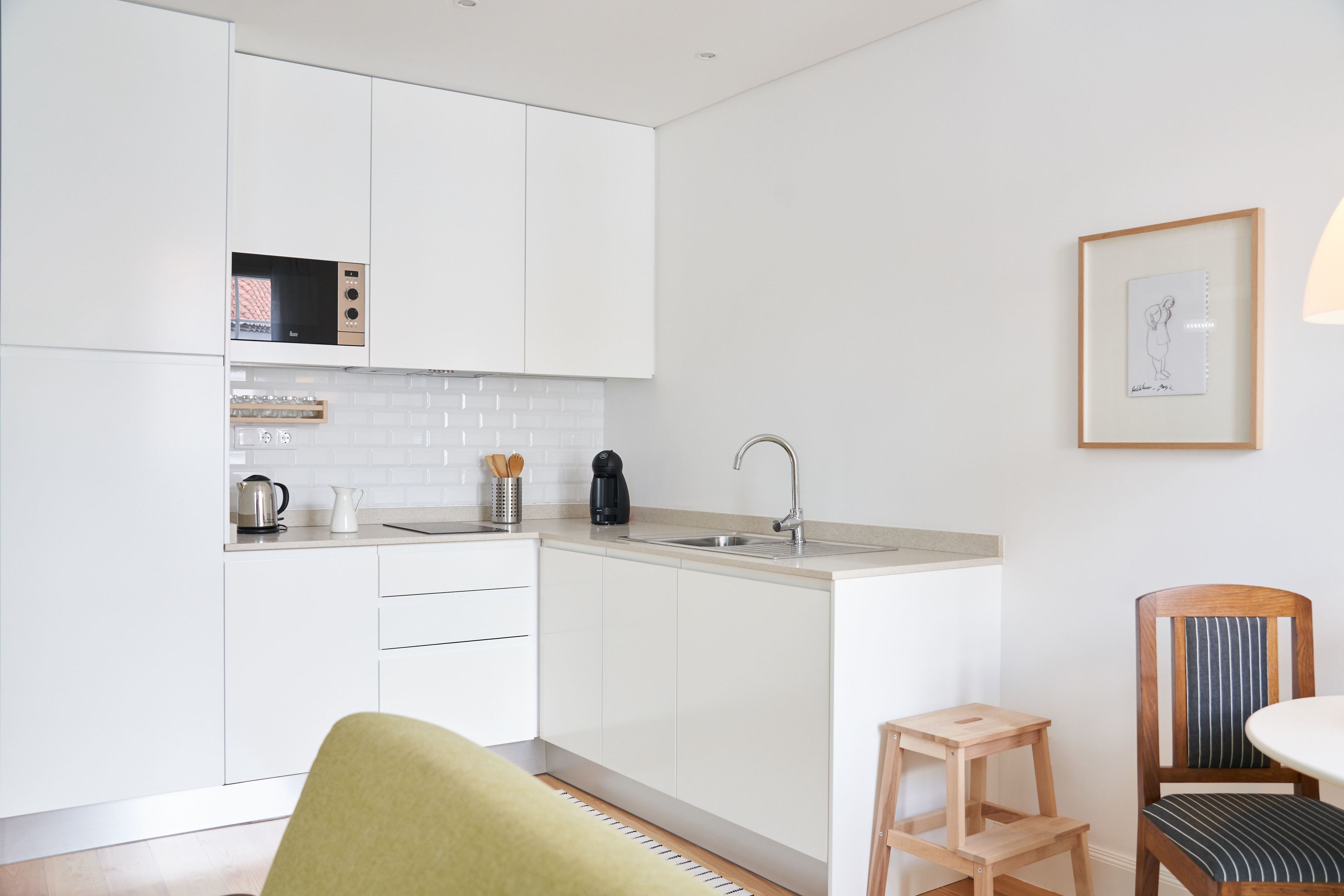Cocina moderna con armarios blancos, microondas integrado y un lavamanos. Hay un cuadro en la pared y una silla de madera con cojín a rayas.